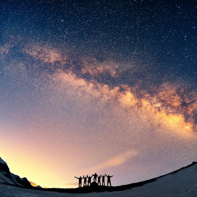 Teamwork and support. A group of people are standing together holding hands against the Milky Way in the mountains.